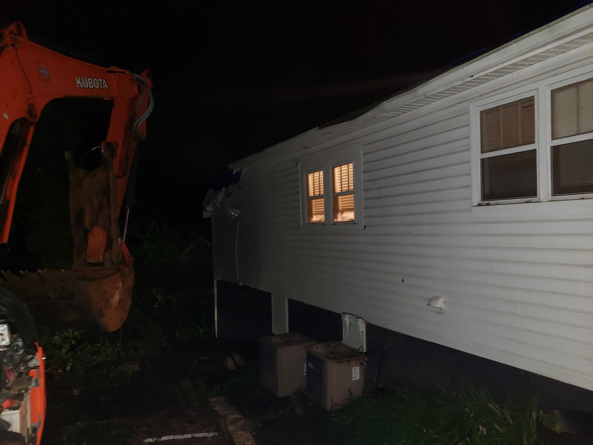 An excavator near a house at night, damaging the siding.