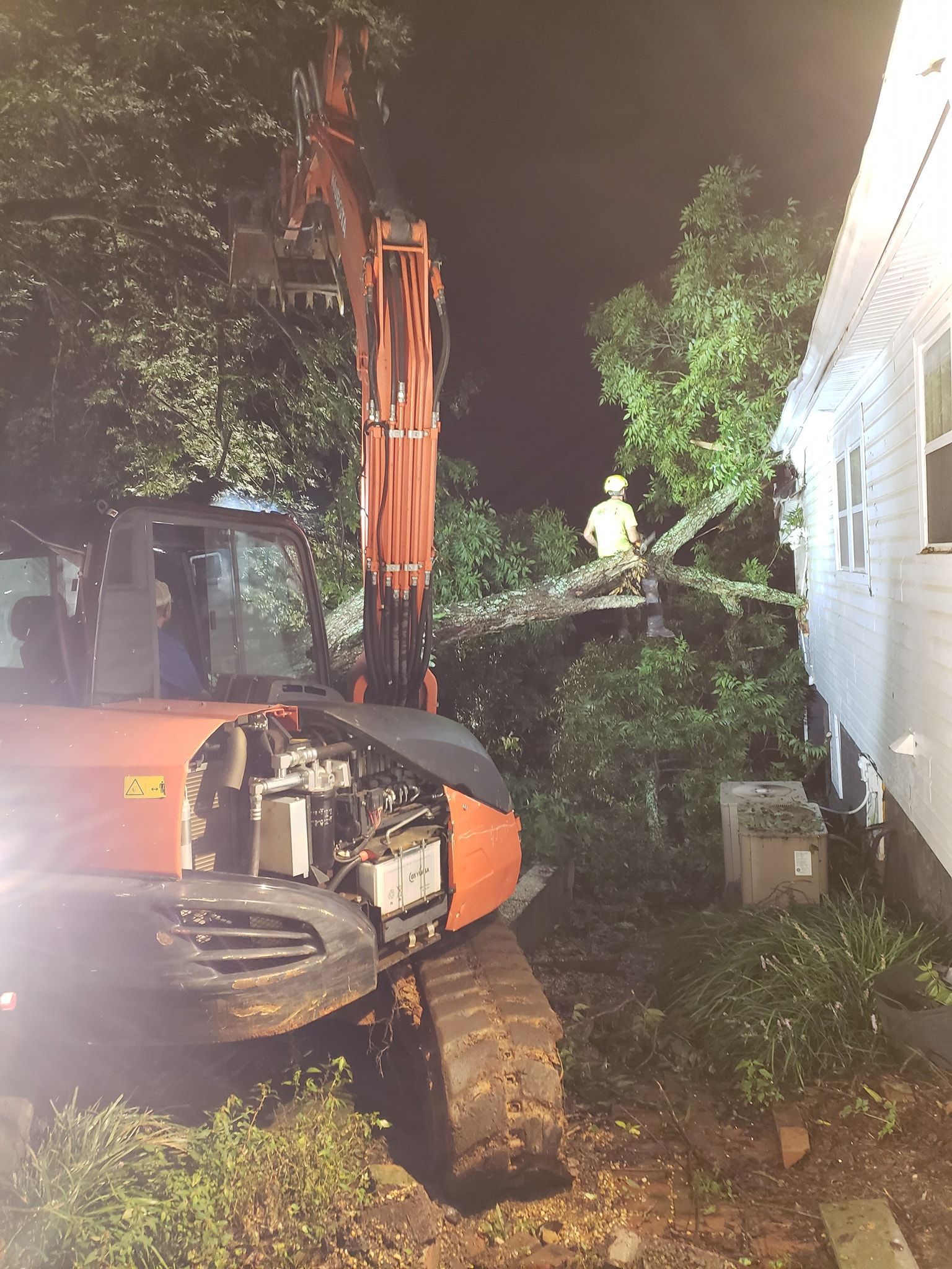 An excavator removing tree branches next to a house at night; a worker in a hard hat is visible.
