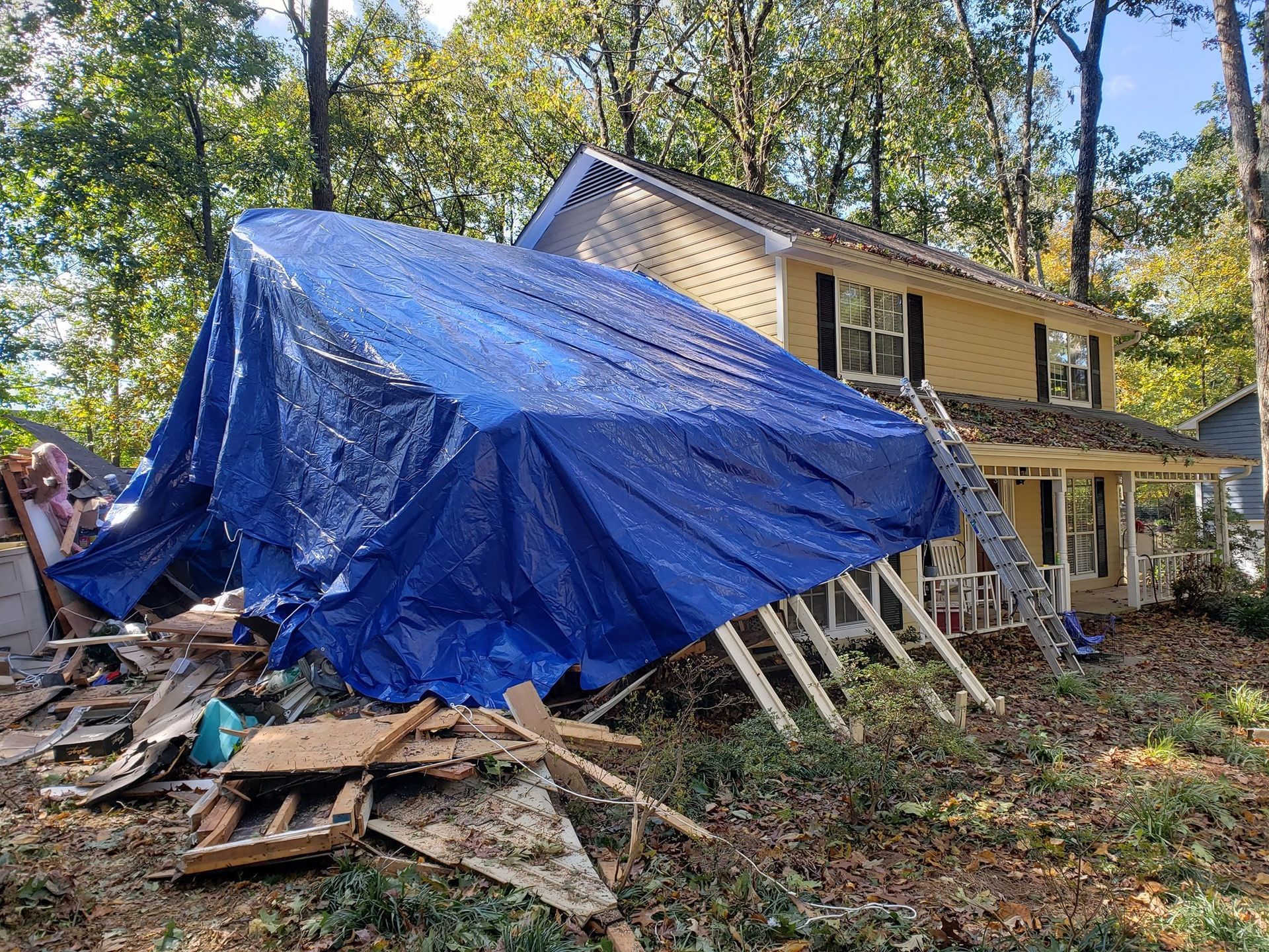 House under blue tarp with debris pile in front. Ladder propped against the side. Surrounded by trees.