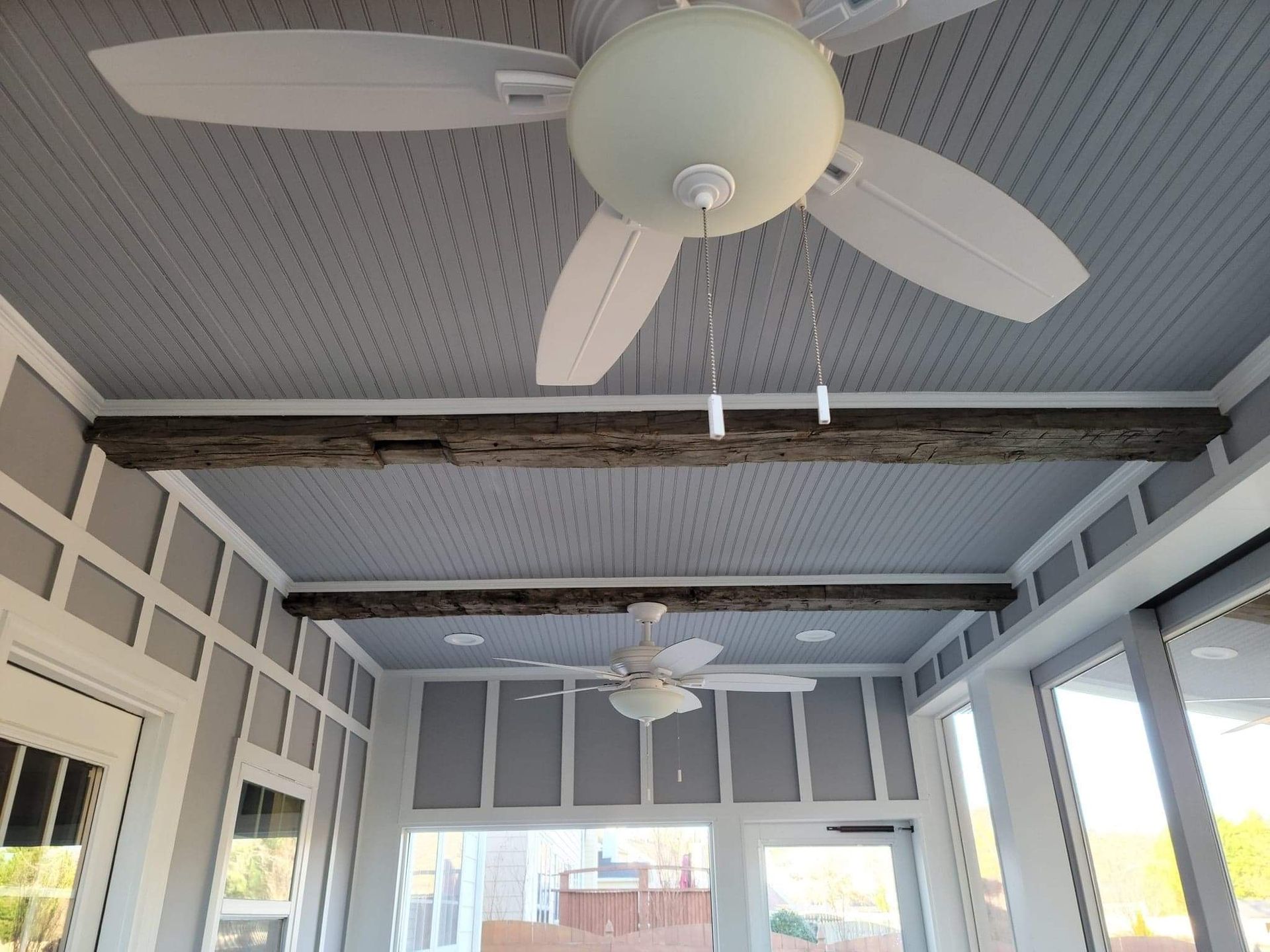 Sunroom with gray ceiling, wooden beams, two ceiling fans, and glass doors.