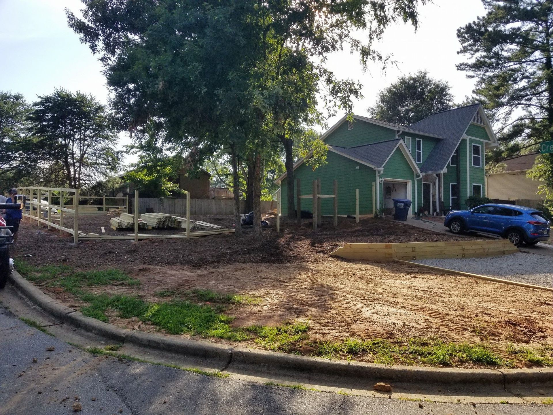 Green house under construction with blue car in driveway; fence and dry dirt lot in the foreground.