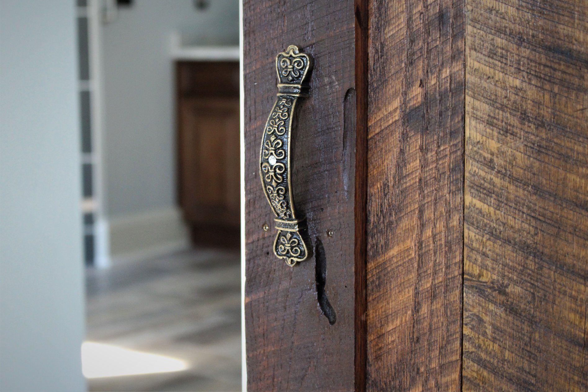 Ornate brass handle on a rustic, dark wooden door. Blurred interior with cabinets visible.