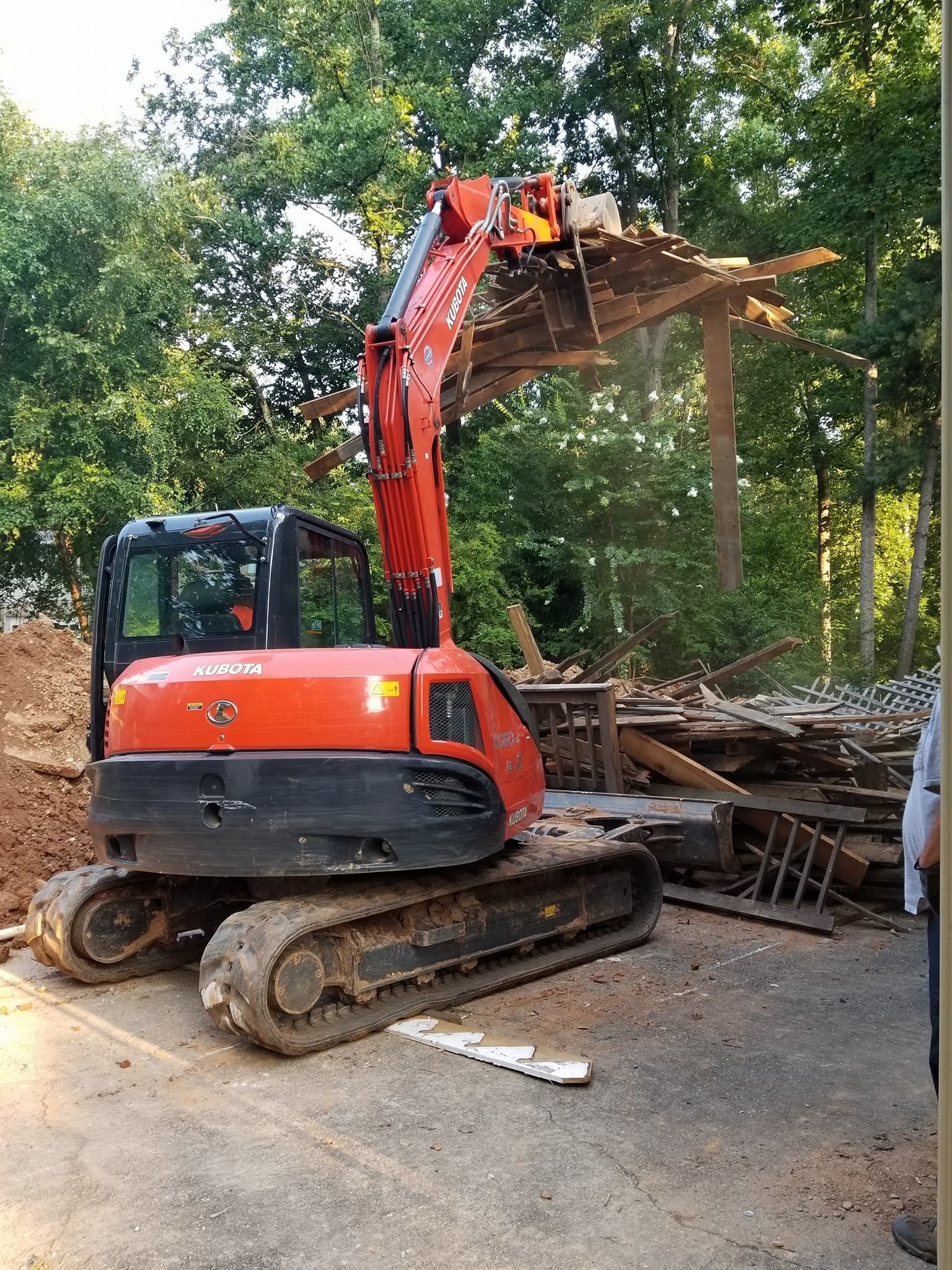 An orange excavator demolishes a wooden structure outdoors amid trees and debris.