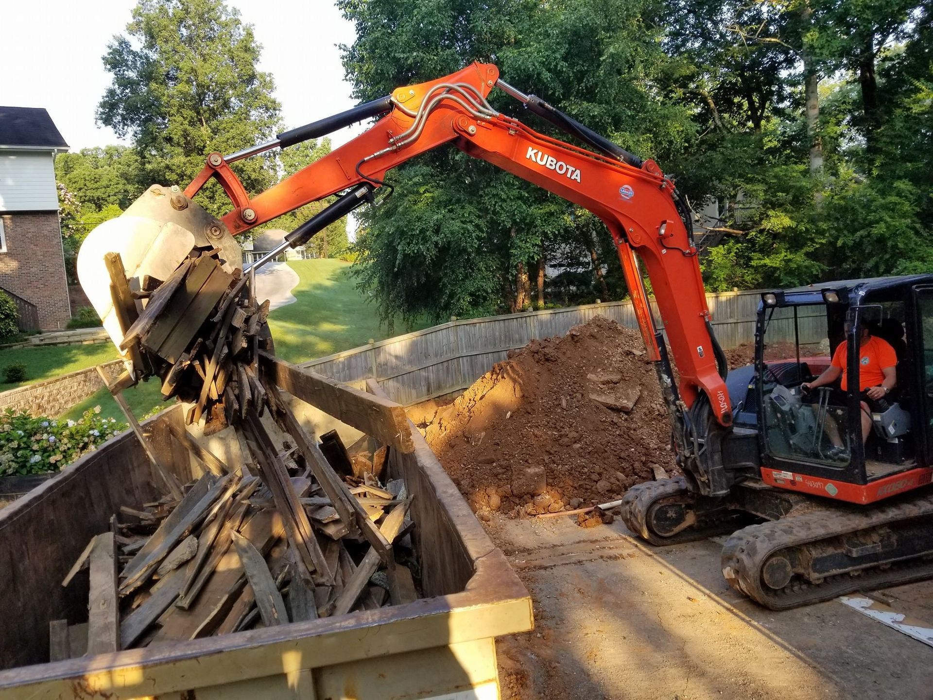 Orange excavator dumping wood debris into a container on a driveway.