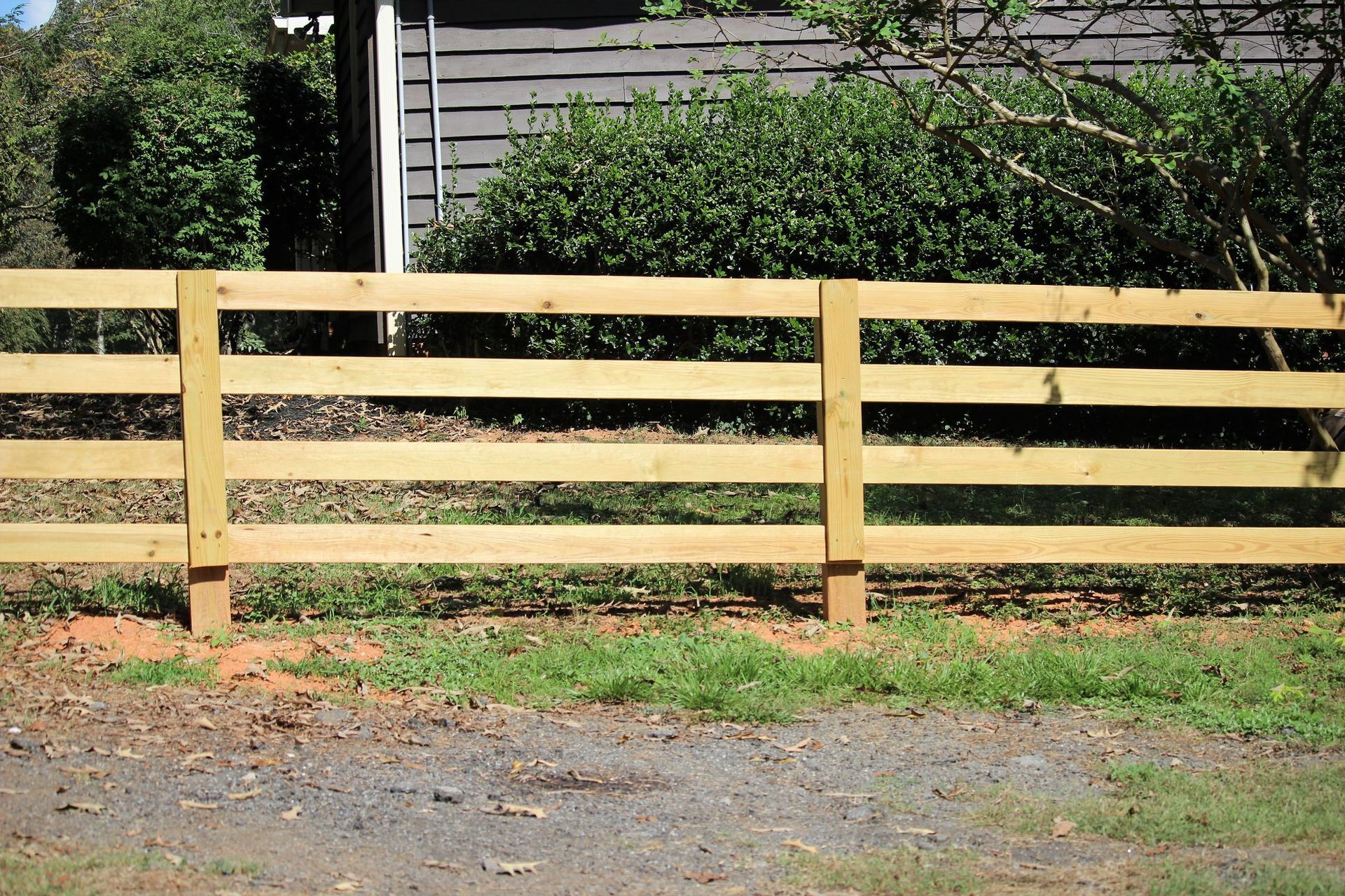 Wooden three-rail fence with posts, in front of a building and bushes.