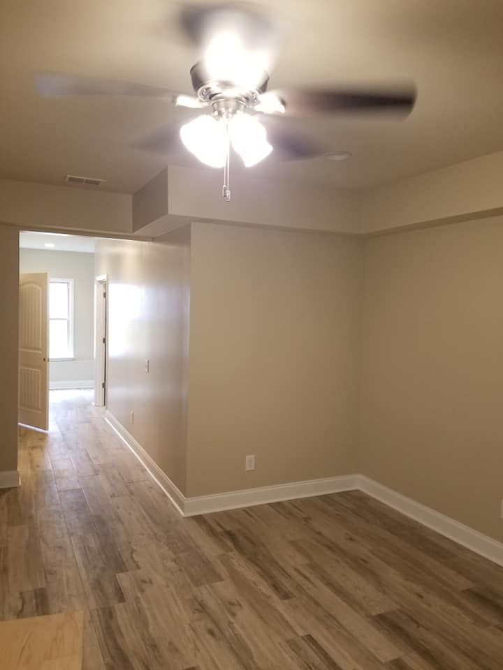 Empty bedroom with beige walls, wood-look flooring, and a ceiling fan; doorway leads to a hallway.