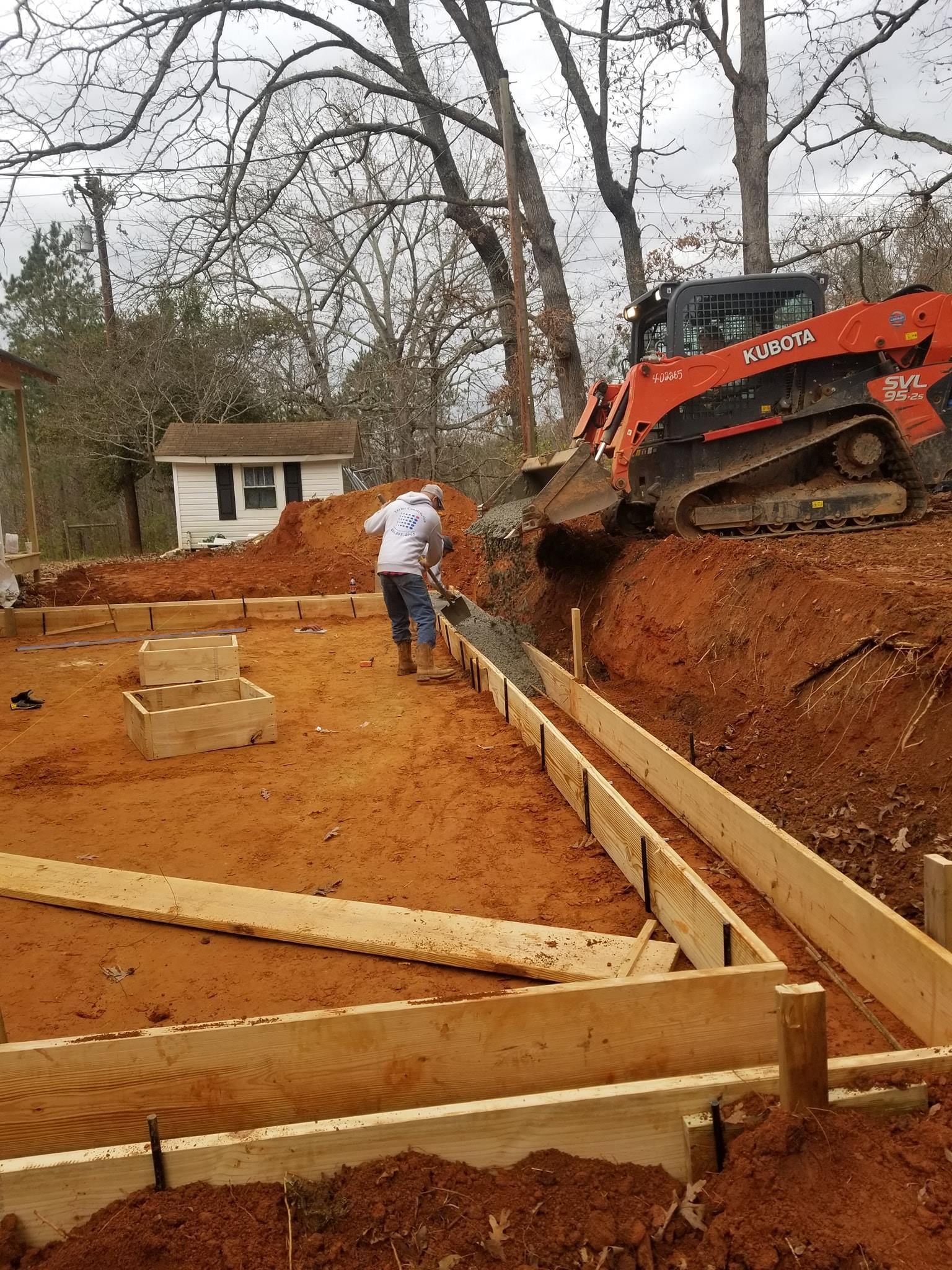 Construction site: Man working on wooden forms, excavator in background, red dirt.