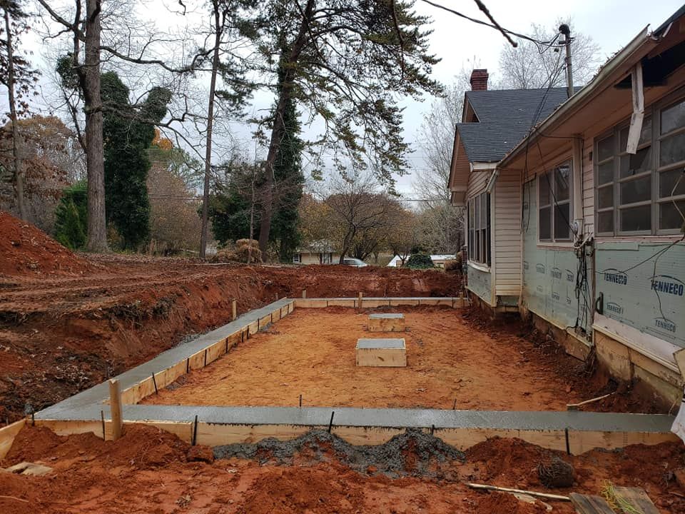 Construction site with new concrete foundation next to a house; dirt, wood, and trees.