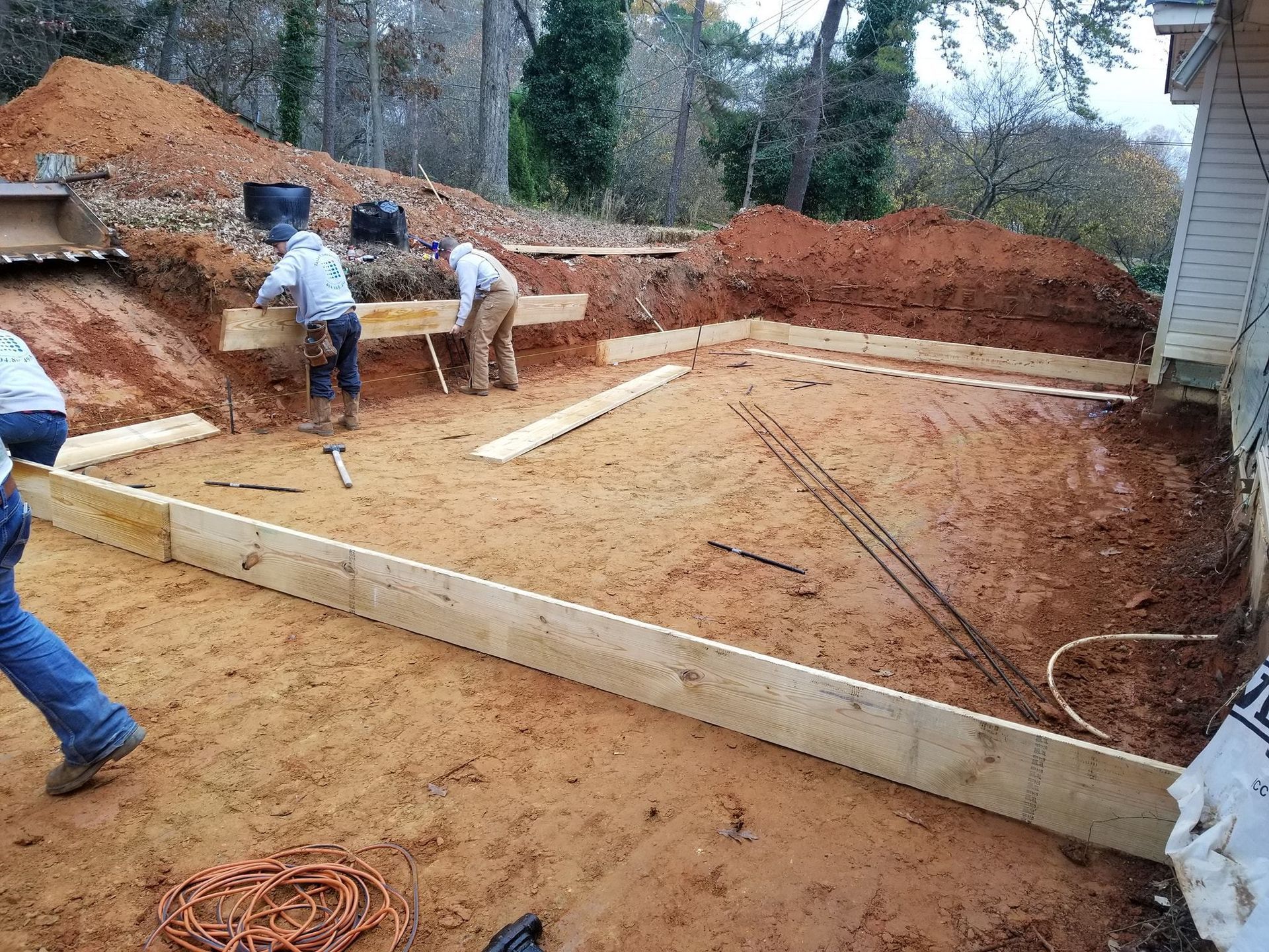 Construction workers building a foundation with wooden forms on a dirt surface, brown soil, and trees in the background.
