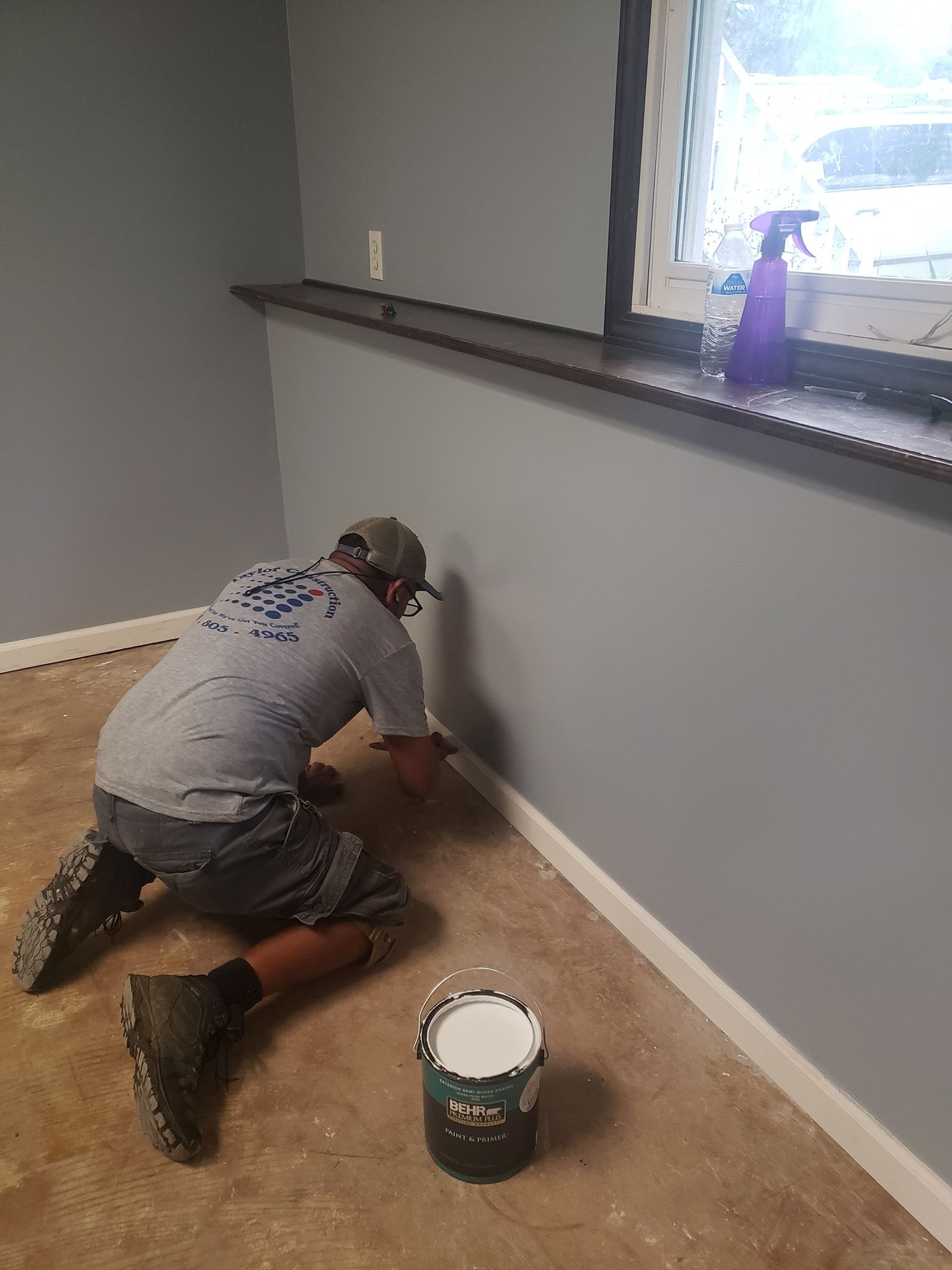 A person painting baseboard in a room with gray walls. A paint can and boot are visible.