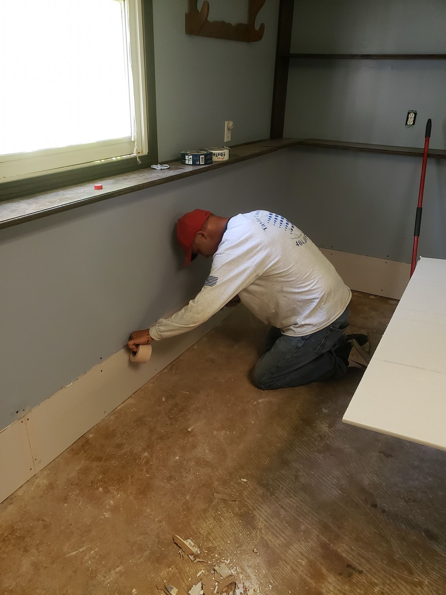 Man in red hat and white shirt installing trim in a room with grey walls and a window.