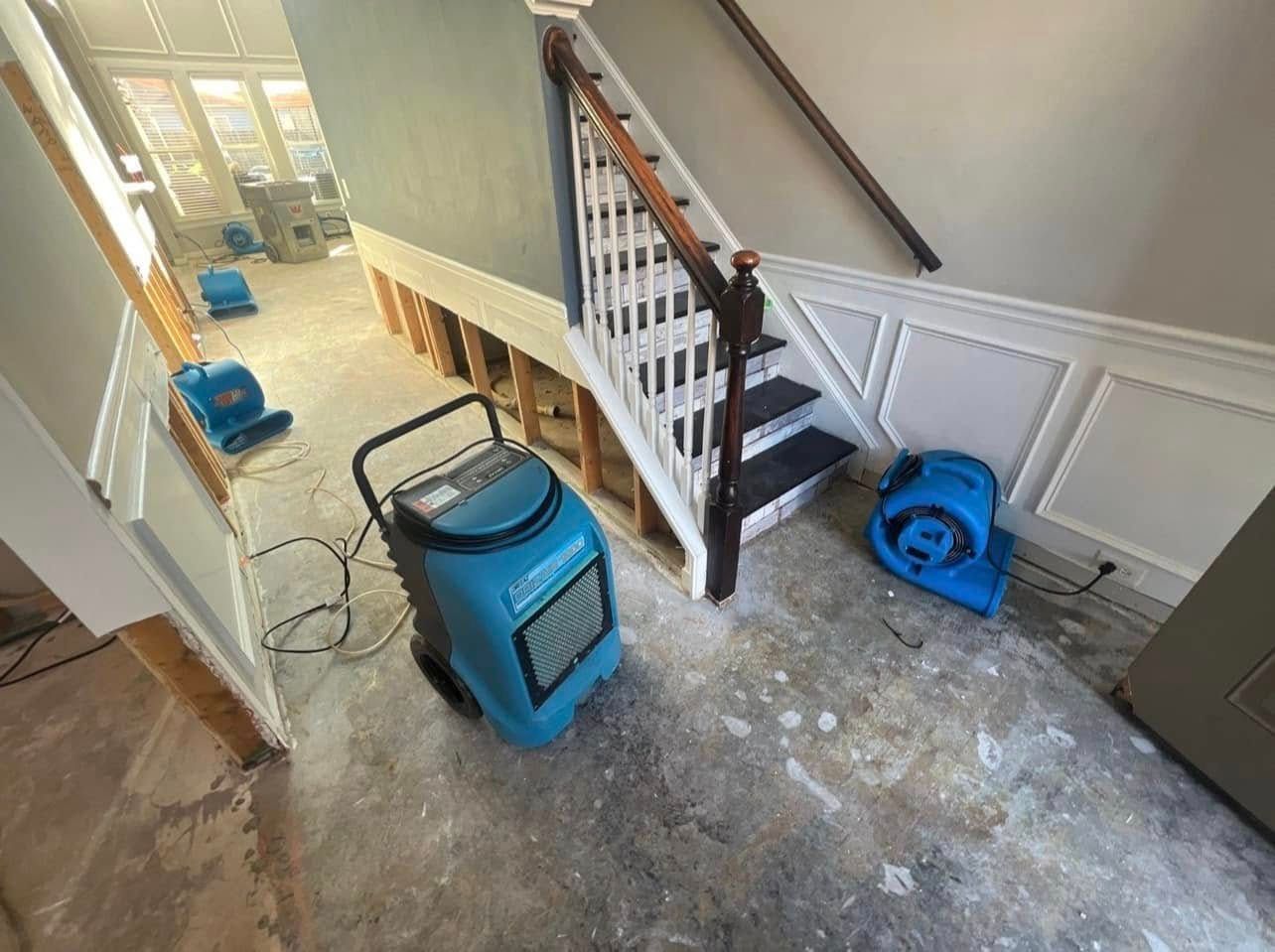 Blue dehumidifiers and fans drying out a flooded entryway, near a staircase with exposed framing.