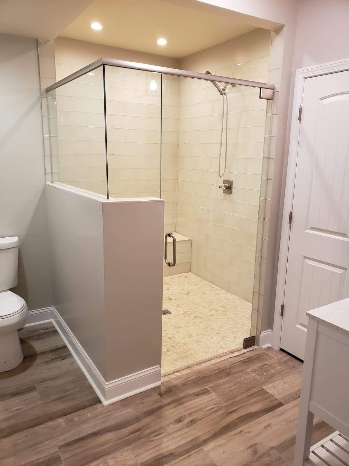 Modern bathroom with glass shower, white walls, and wood-look flooring. Toilet and sink visible.