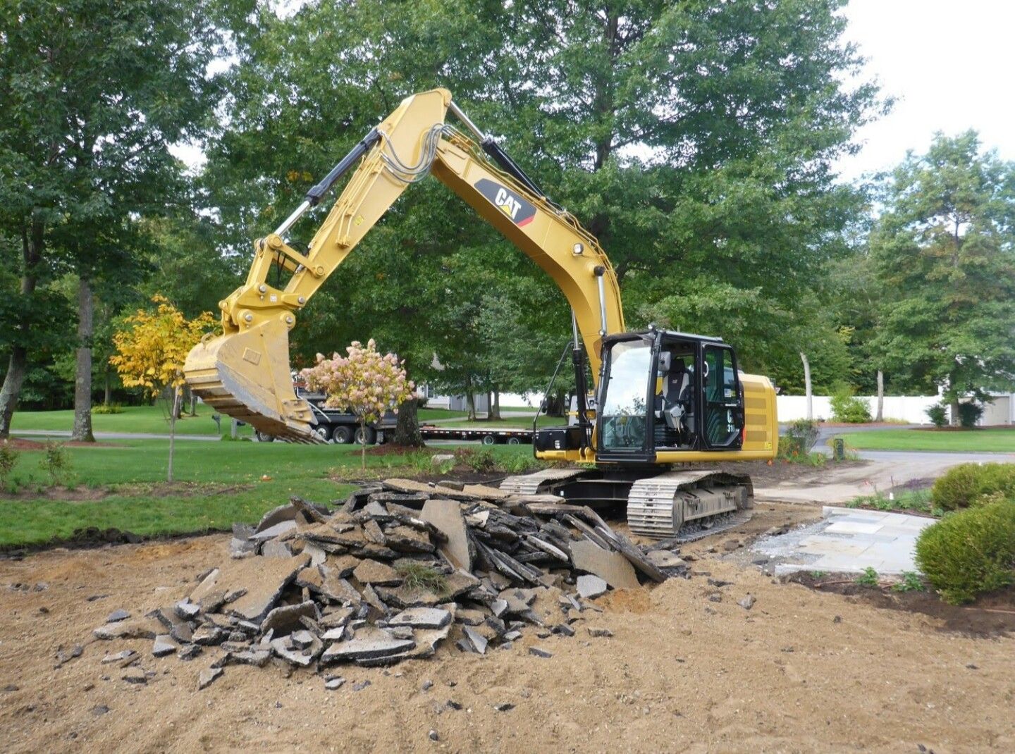 Yellow excavator removing asphalt debris on a residential driveway, trees in background.