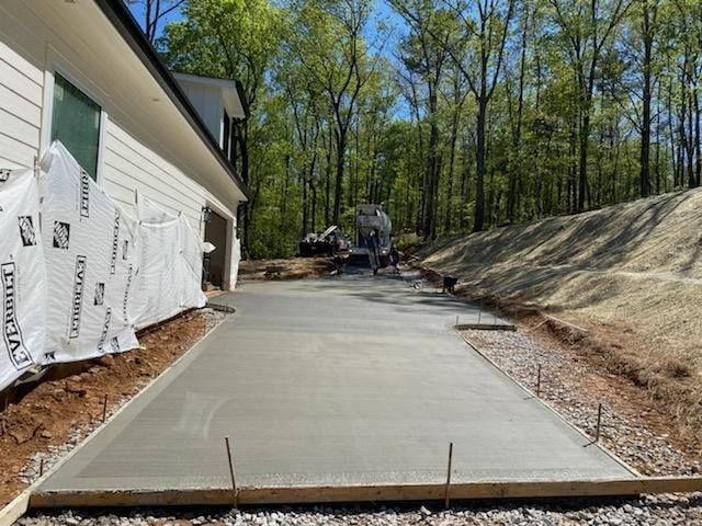Newly poured concrete driveway next to a house and wooded hillside; cement truck in the background.