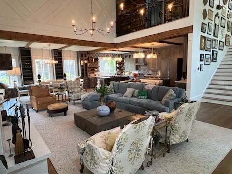 Spacious living room with exposed beams, gray couches, floral chairs, and a large fireplace, leading to kitchen.