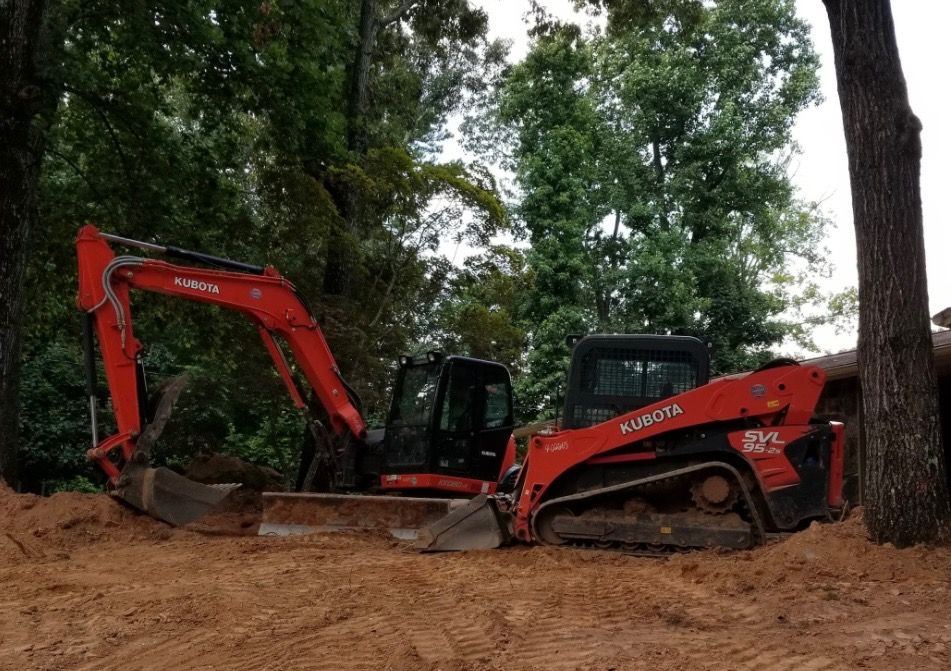 Red Kubota excavator and skid steer working on a dirt pile near trees.