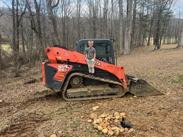 Boy stands on an orange Kubota skid steer. Trees and a field are in the background.