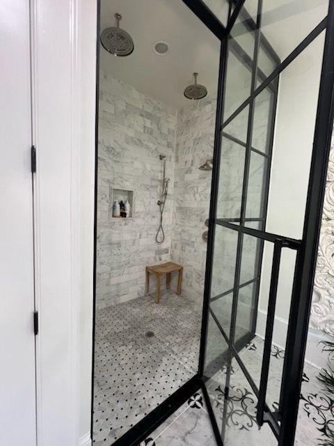 Shower with black framed glass door, white marble tile walls, and mosaic floor tiles, wood stool.