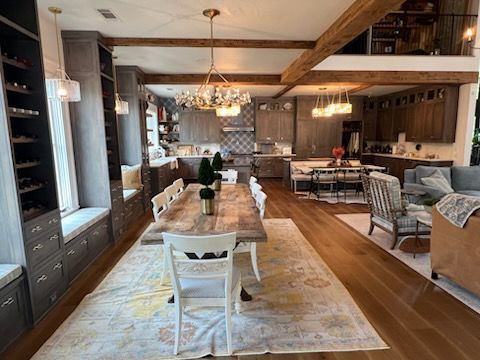 Open-concept dining area with a long table, adjacent to a kitchen, and a living room. Wooden beams and hardwood floors.