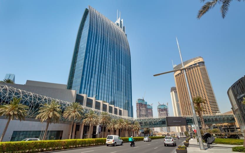 Tall, blue glass skyscraper with curved top in Dubai, palm trees and road in foreground.