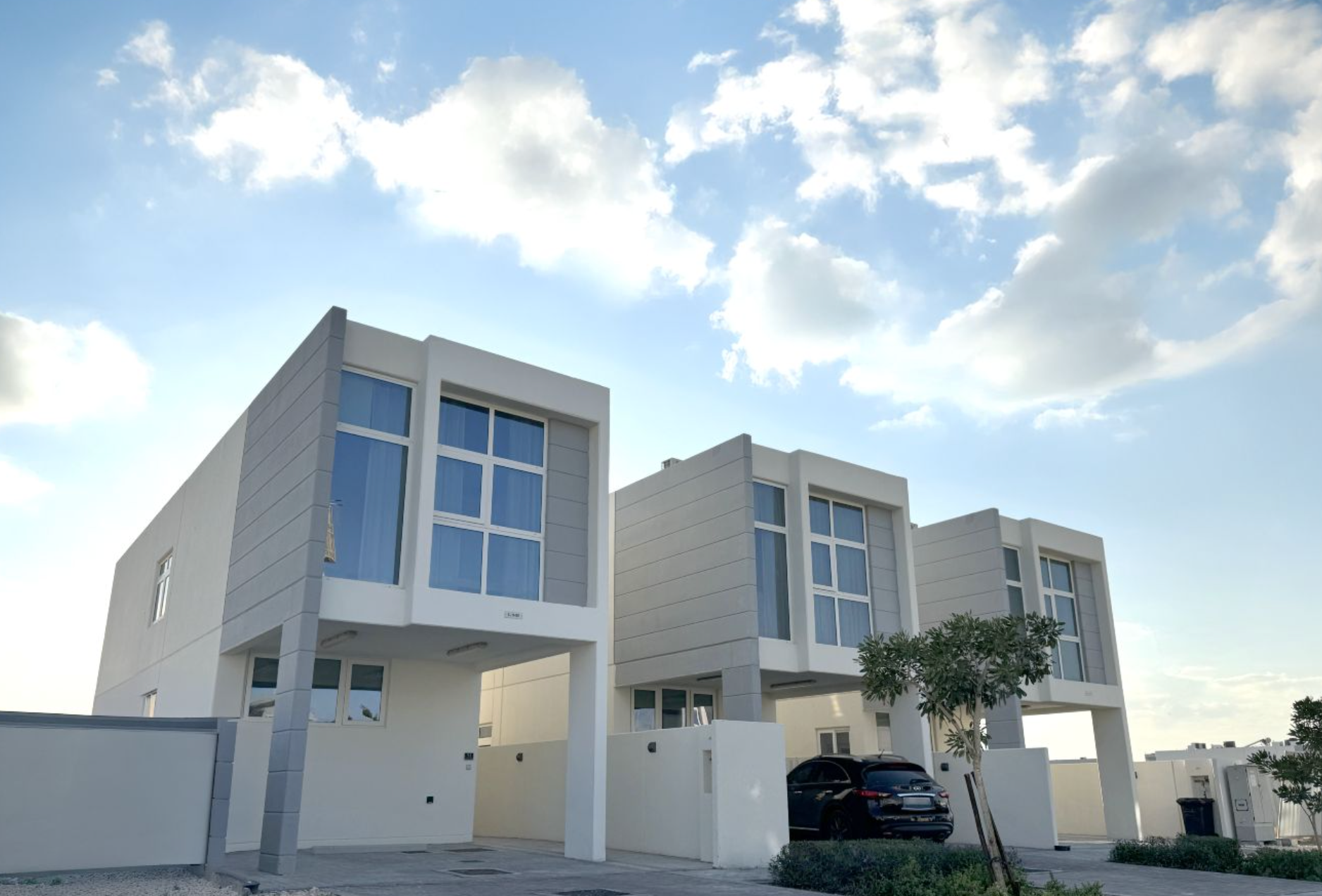 Three modern, white two-story houses with large windows under a blue sky with clouds; a car is parked in the driveway.