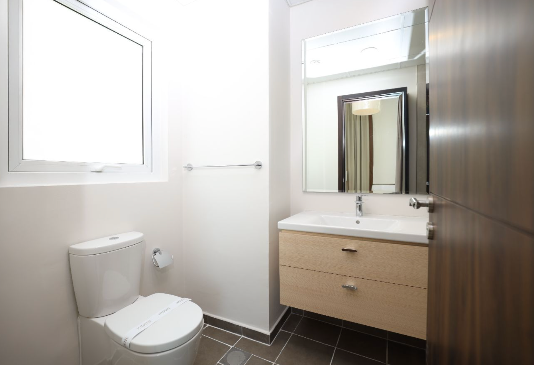 A modern bathroom with a toilet, sink, mirror, and a window. Light wood vanity, white walls.