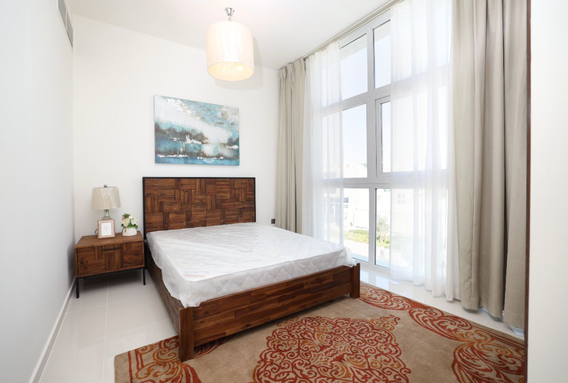 Bedroom with a wooden bed, nightstand, art, and rug. Window with sheer curtains.
