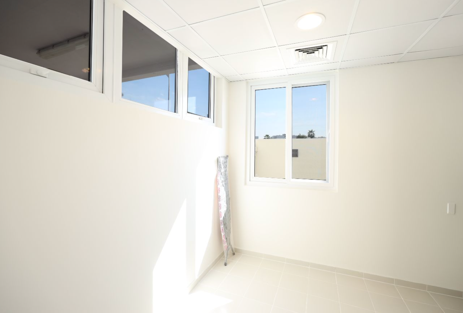 Empty, white-walled room with a window letting in sunlight. An ironing board leans against a wall.