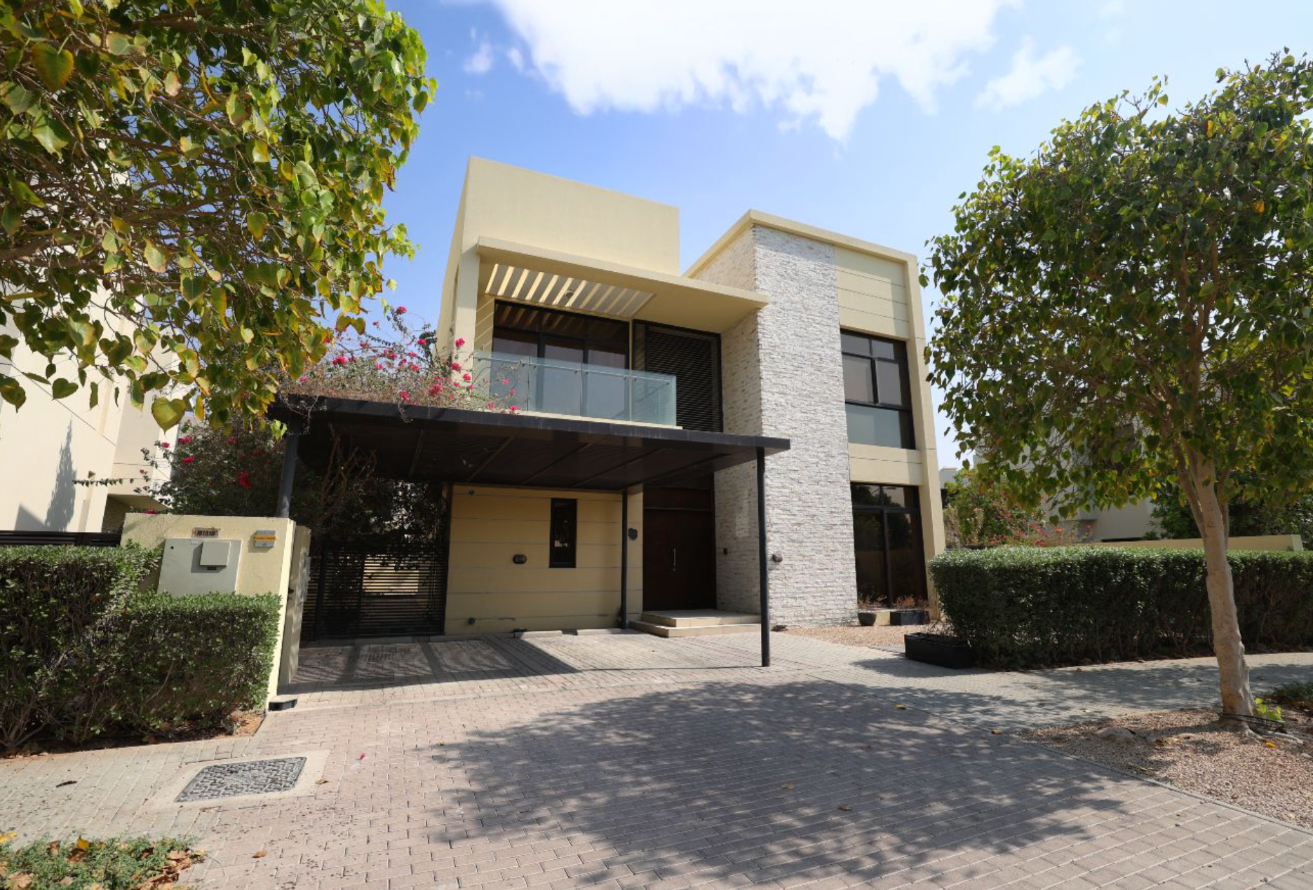Modern two-story house with beige and stone facade, carport, and manicured landscaping under a blue sky.