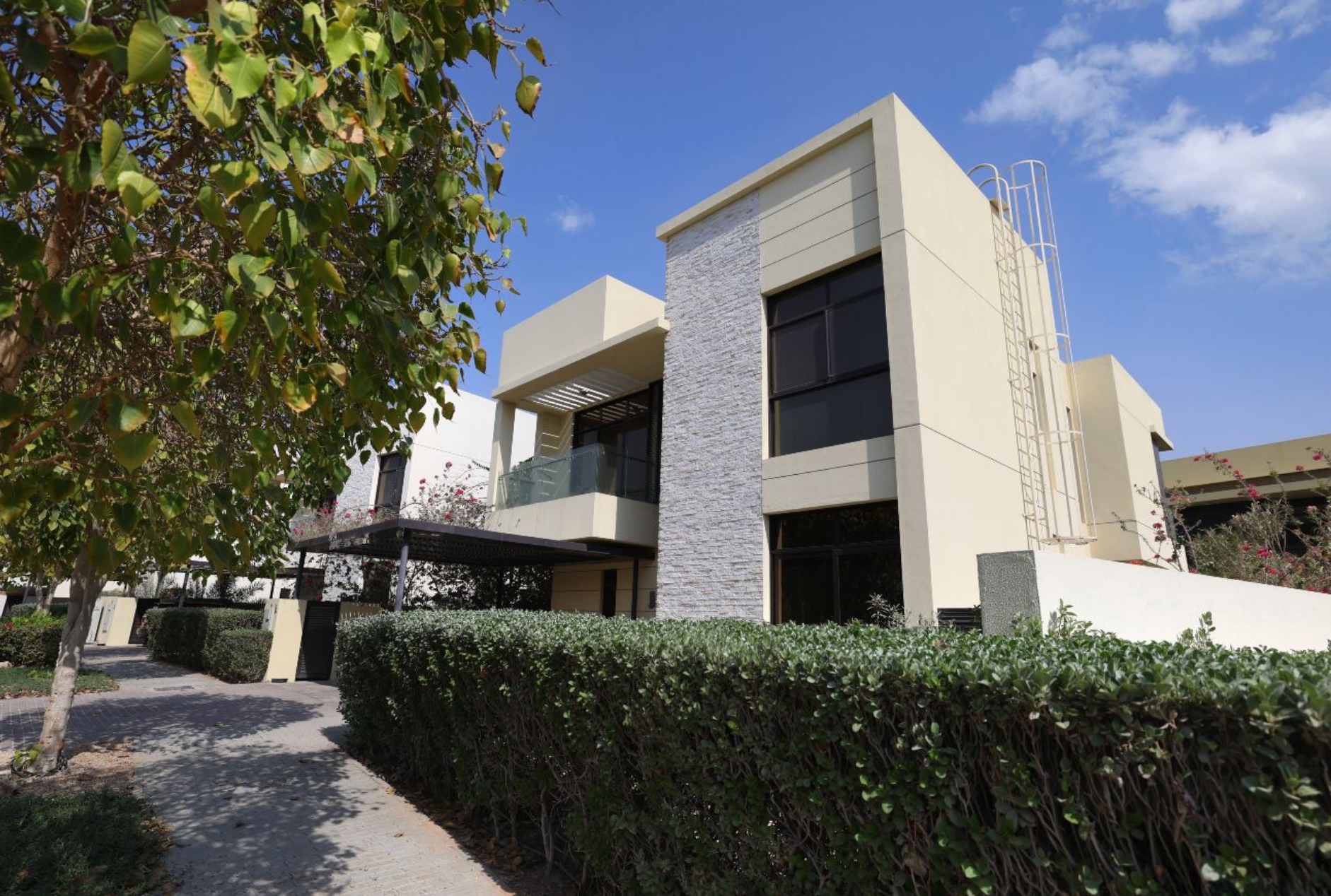 Modern two-story house with tan walls, stone accents, and glass balconies. Green hedge and tree in front under a blue sky.