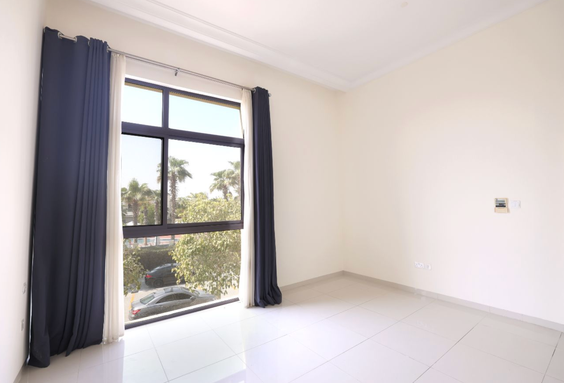 Empty room with large window, dark curtains, white walls, and tiled floor; exterior view.