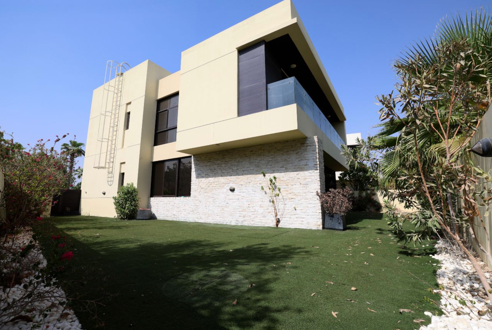 Modern two-story house with beige walls, large windows, and a stone facade, surrounded by a green lawn and bushes.