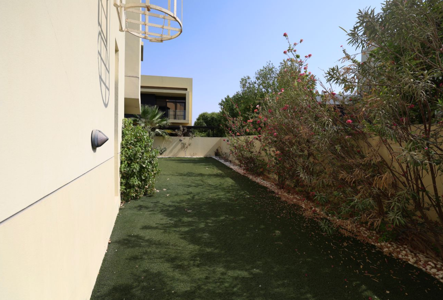 A narrow backyard with green turf, bushes, and a basketball hoop against a beige wall.