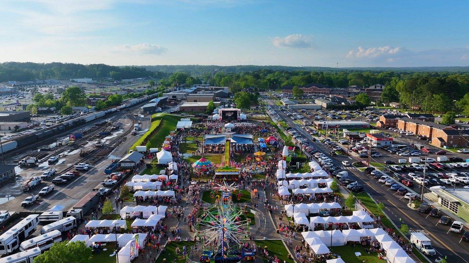 Aerial view of a crowded outdoor festival with white vendor tents, a fairground, and surrounding parking lots.