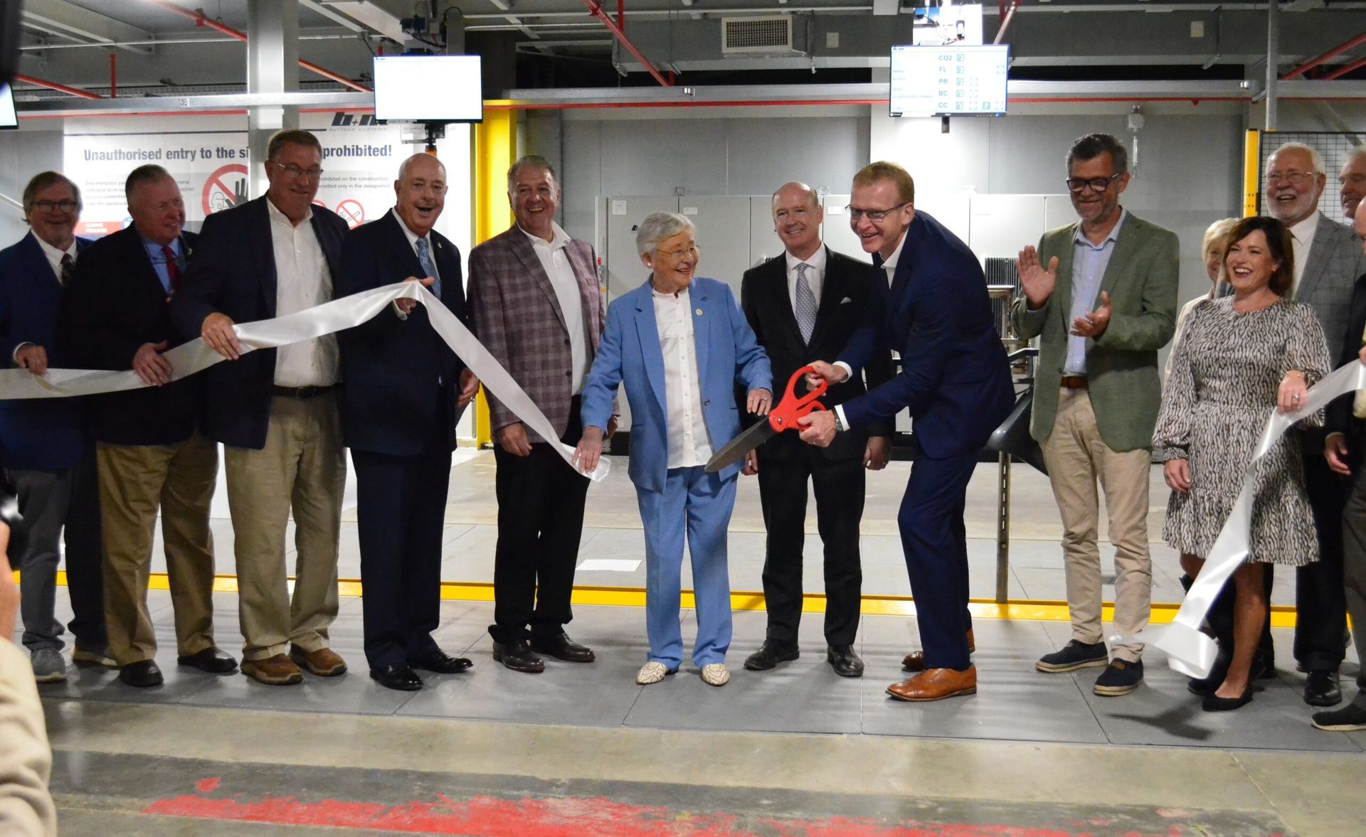 A group of people are standing around a Gov. Kay Ivey cutting a ribbon.