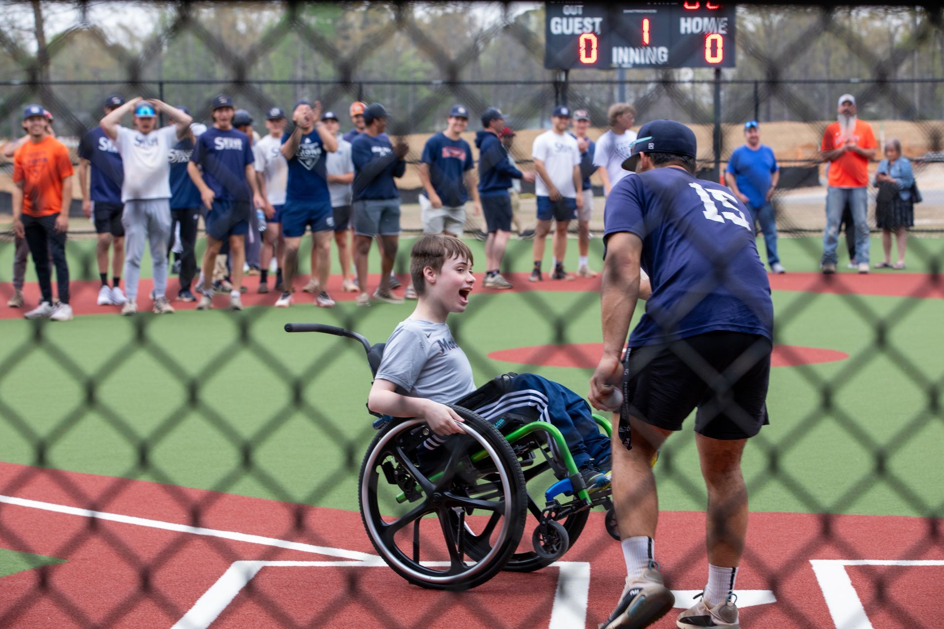 A man in a wheelchair is playing baseball with a group of people behind a chain link fence.