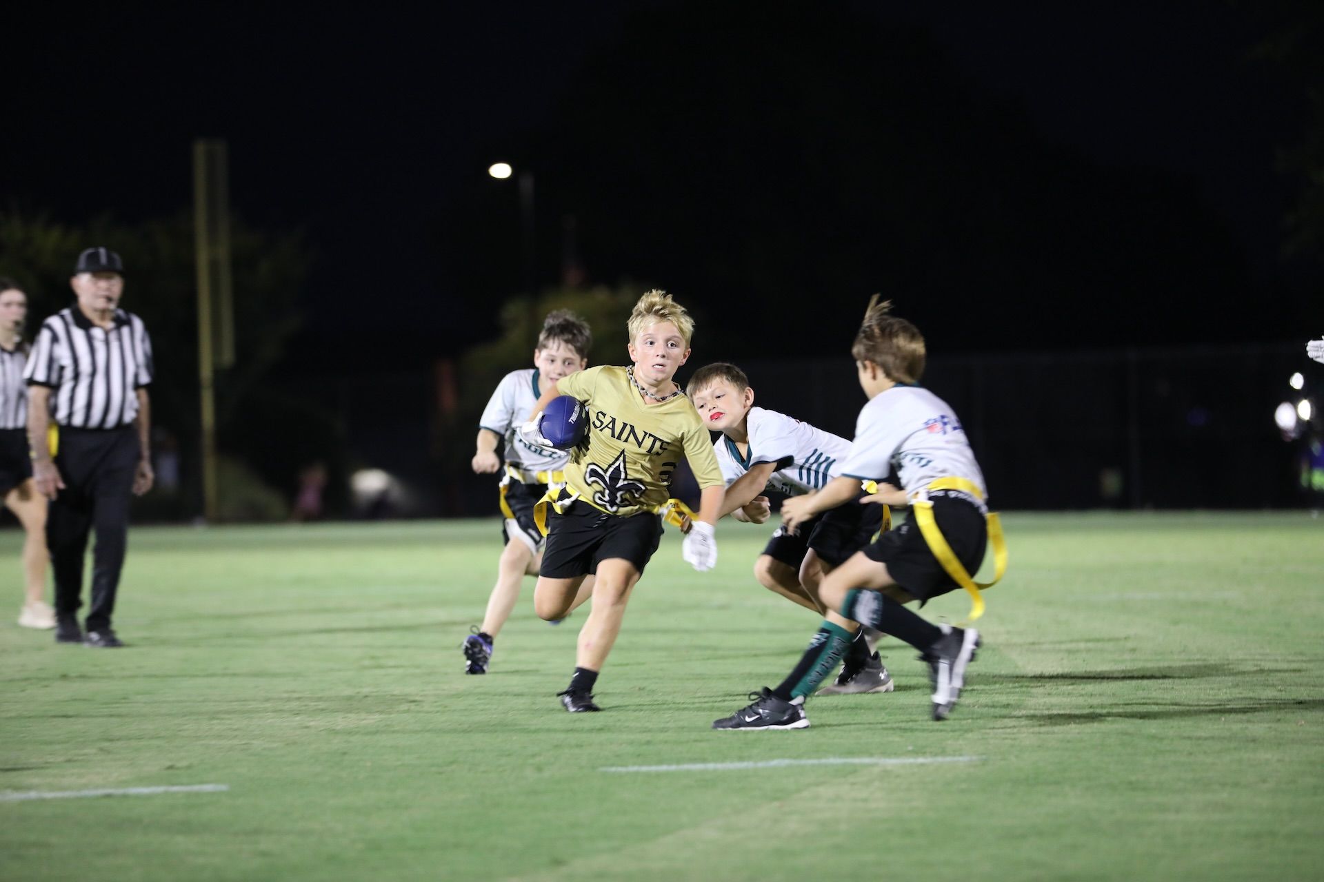 A youth flag football player in a tan jersey runs with the ball as two opponents attempt to pull their flags at night.