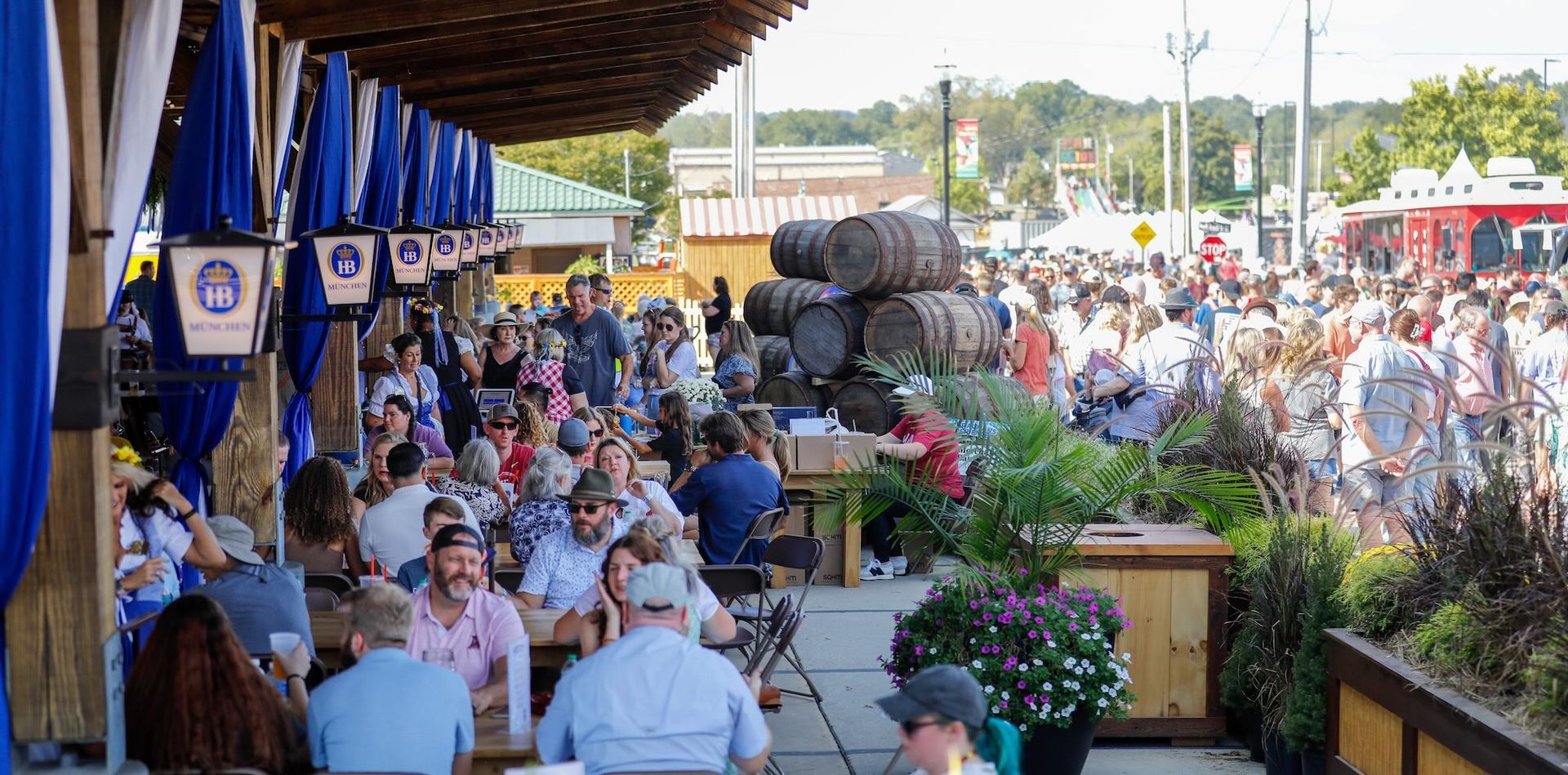 Hofbräuhaus outdoor beer garden with seated people, blue umbrellas, flower boxes, and stacked barrels along a covered patio