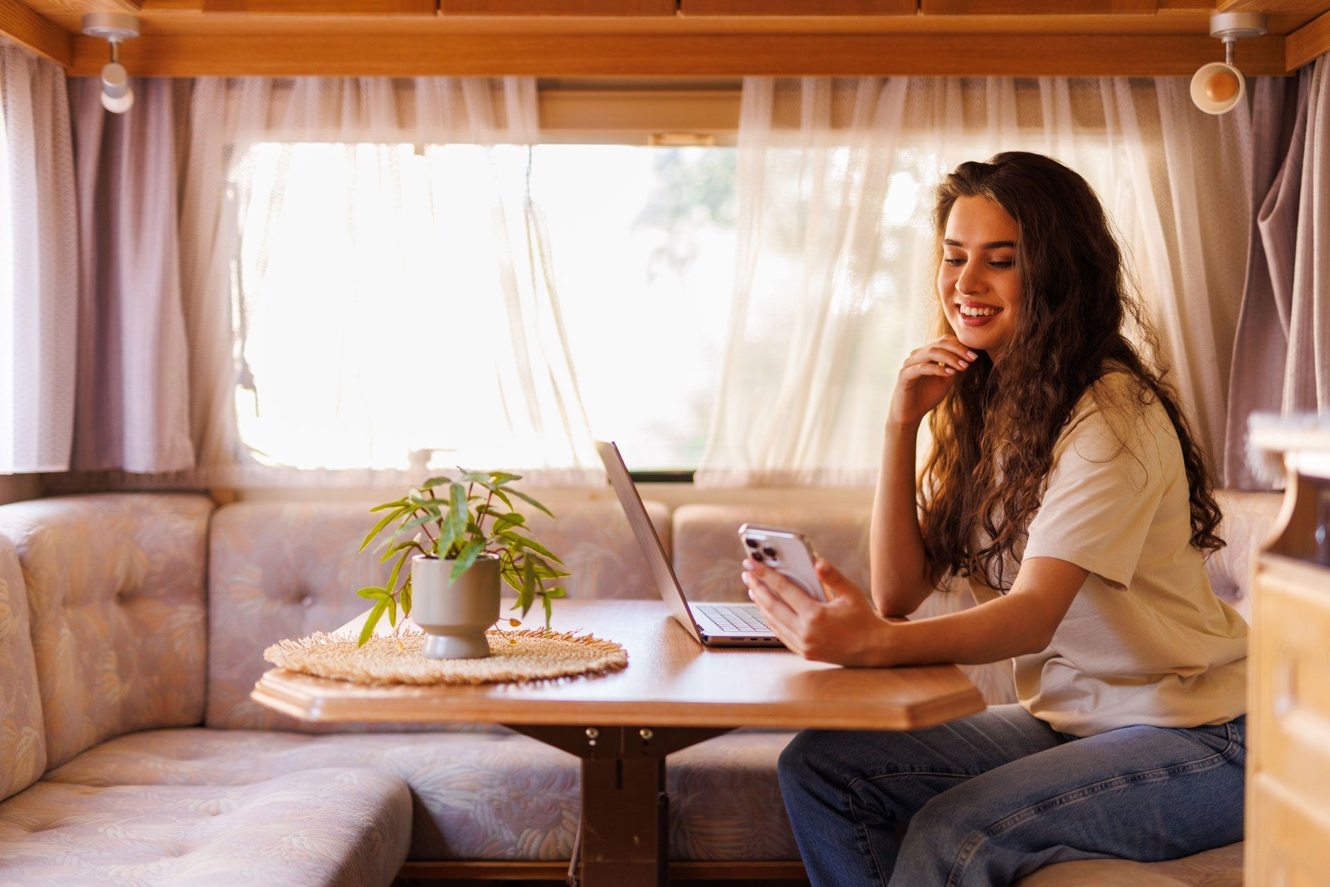 Woman using laptop and phone, smiling inside an RV with a small plant on the table.