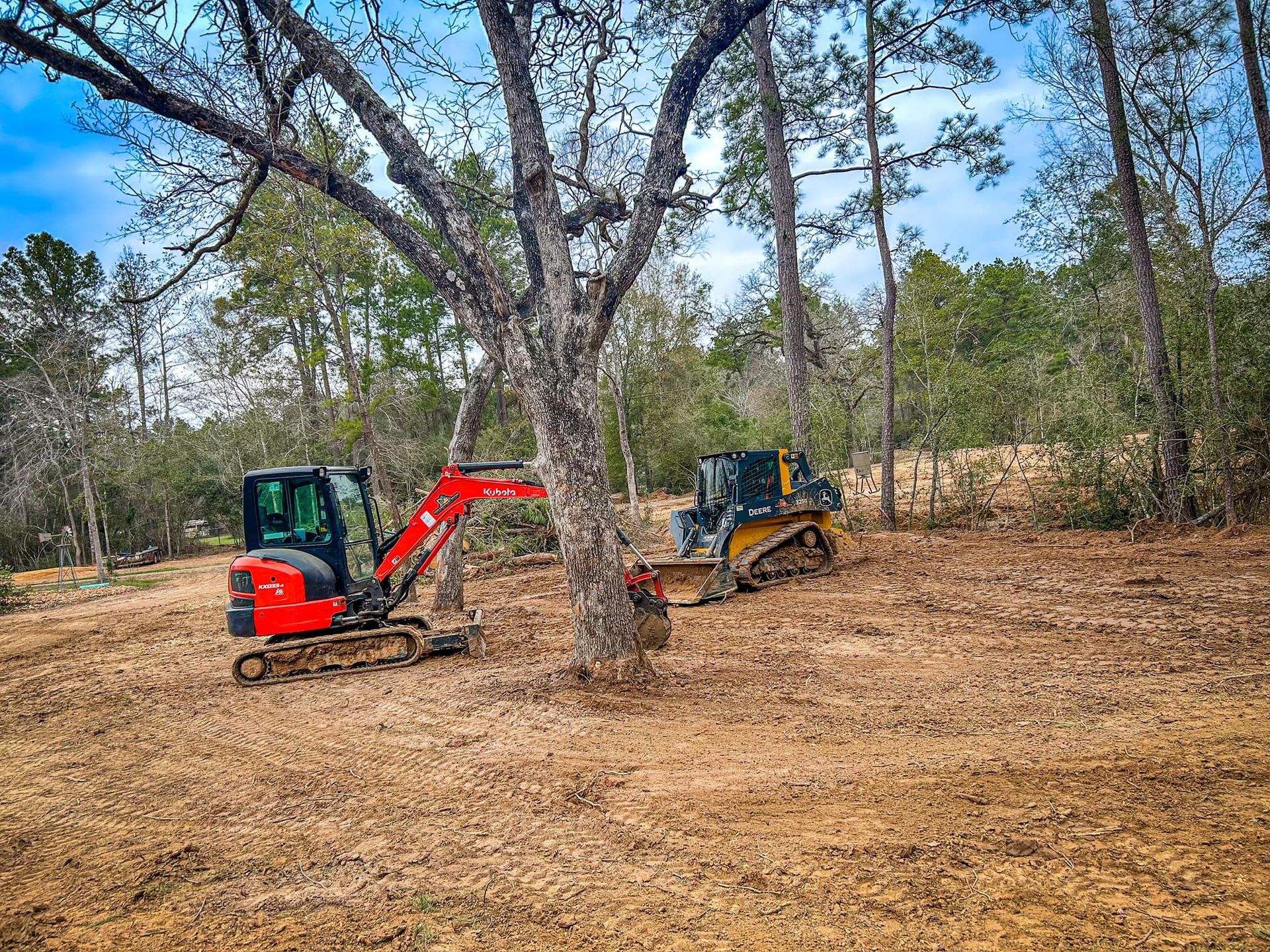 An excavator and a dozer clearing land near trees in a wooded area, with a brown leaf-covered ground.