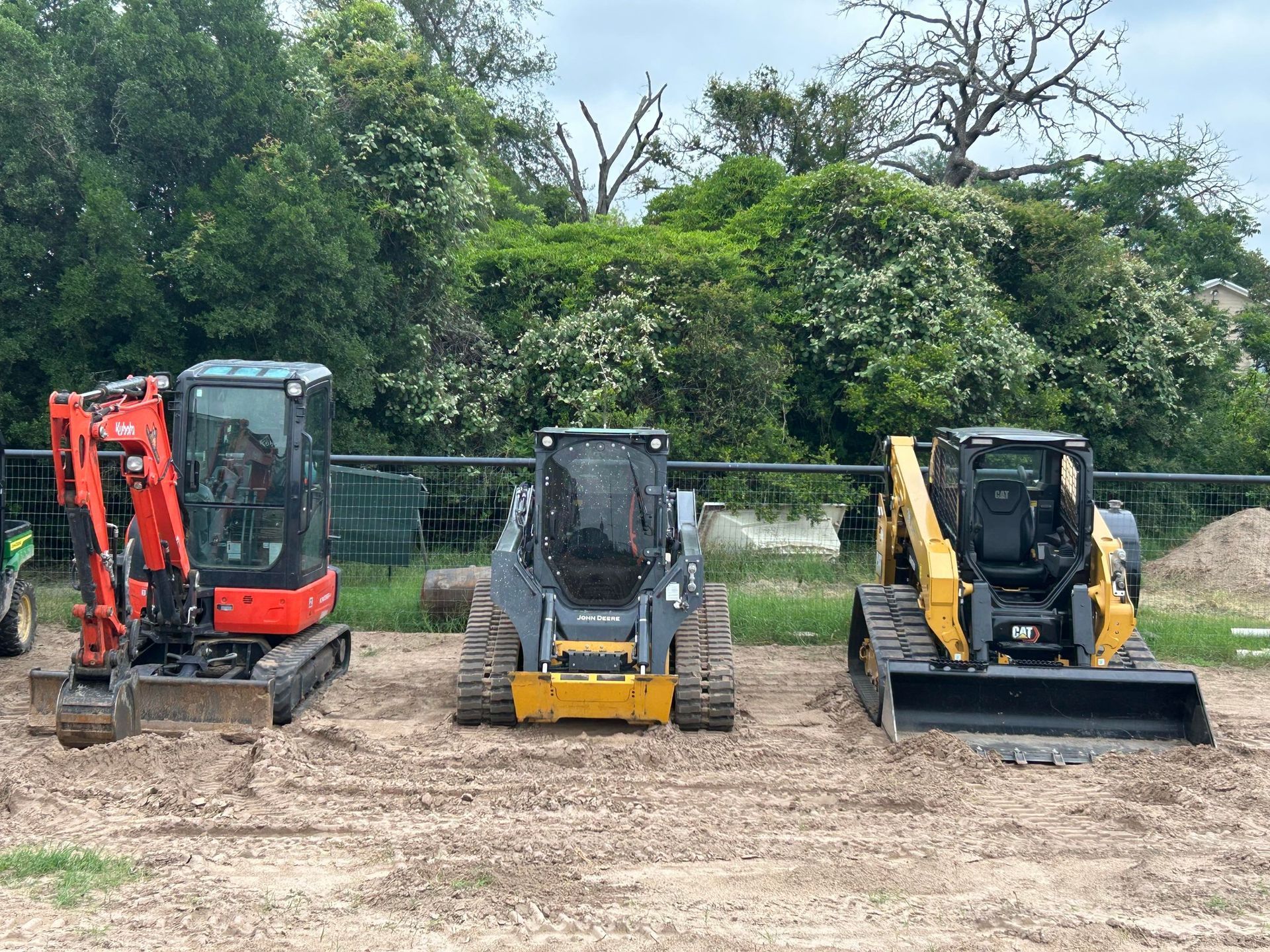 Three pieces of construction equipment parked side-by-side outdoors: an orange excavator, a gray skid steer, and a yellow track loader.