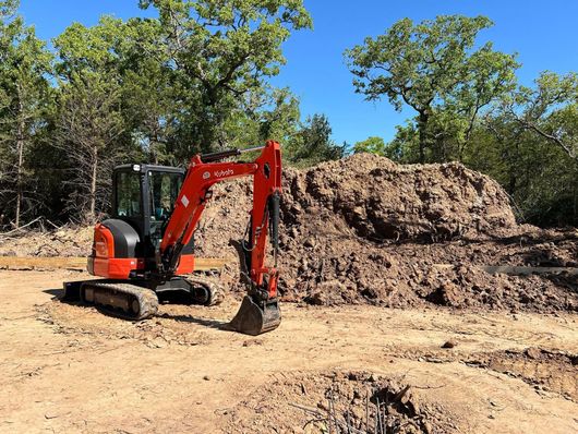 Small orange excavator in a dirt lot with a large pile of dirt in the background and trees under a blue sky.