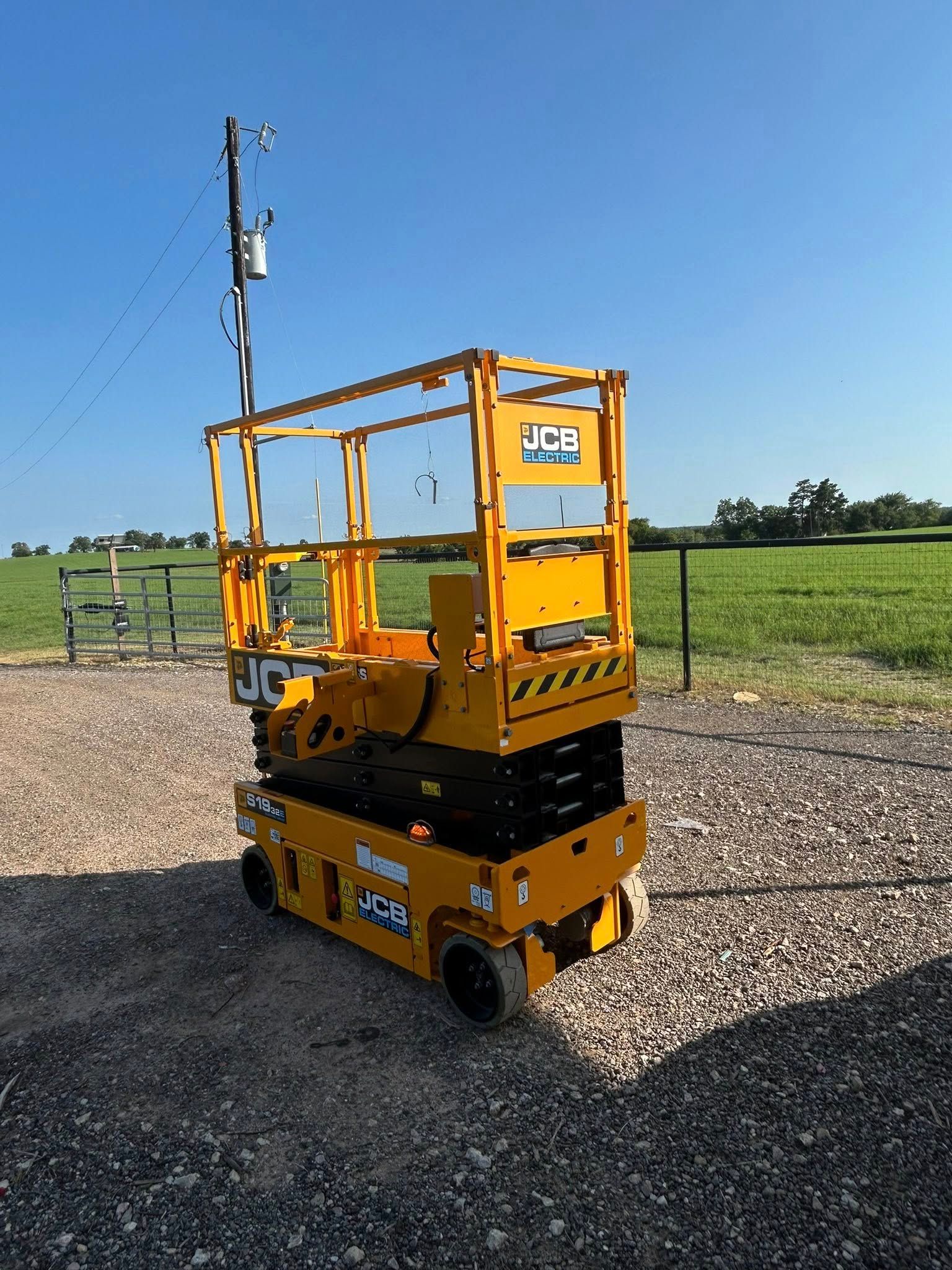 Yellow JCB scissor lift outdoors on gravel in front of a grassy field and blue sky.