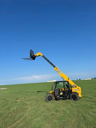 Yellow telehandler with extended boom and forks on a grassy field under a blue sky.