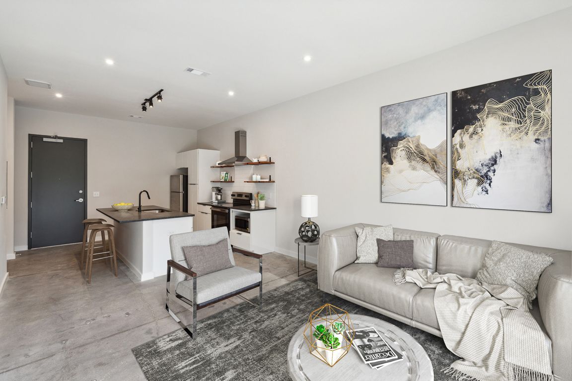 Modern living room with kitchen, gray walls, leather sofa, artwork, and concrete floor.