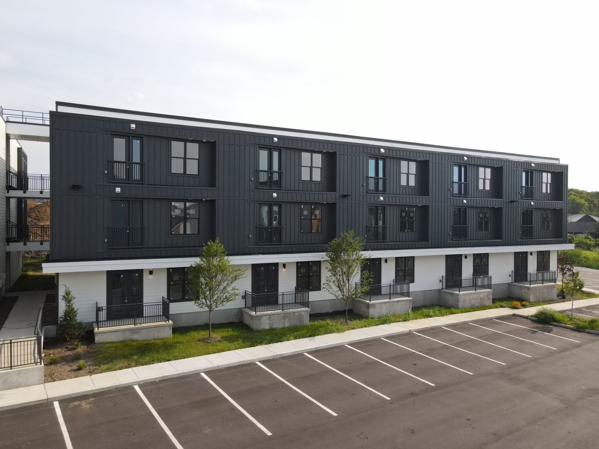 Modern, dark-gray multi-unit building with parking lot. Ground floor has white walls and black doors; upper floors are dark metal.