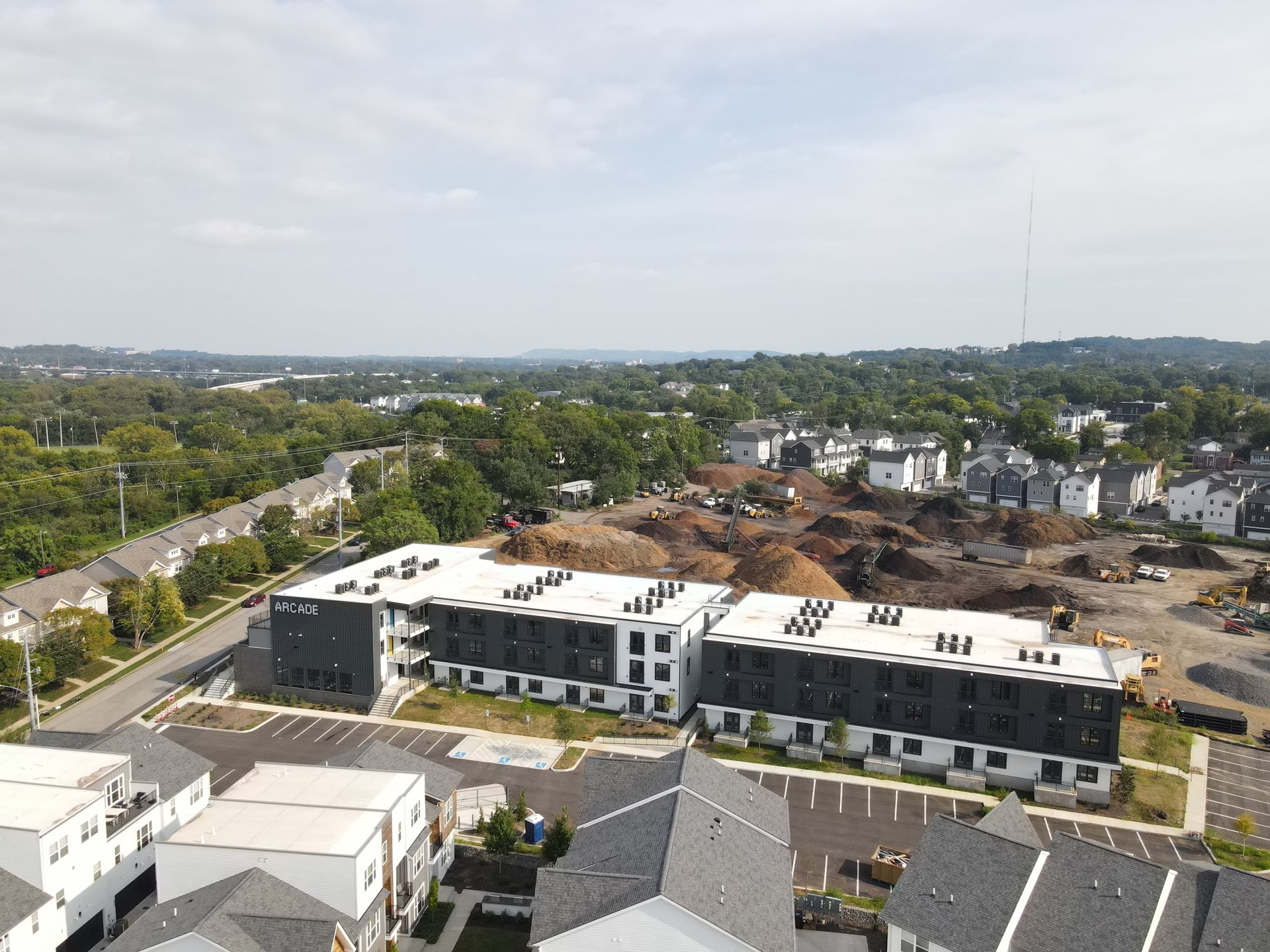 Aerial view of apartment buildings and construction site under a blue sky, with surrounding trees and a cityscape.