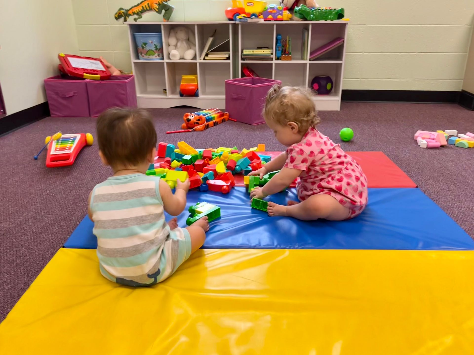Two cute babies play with toys together on a mat in the nursery room