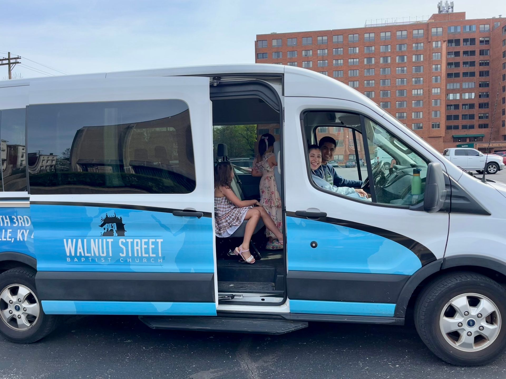 A van labeled Walnut Street Baptist Church. A young man is driving and a young woman is next to him. Kids are in the van.